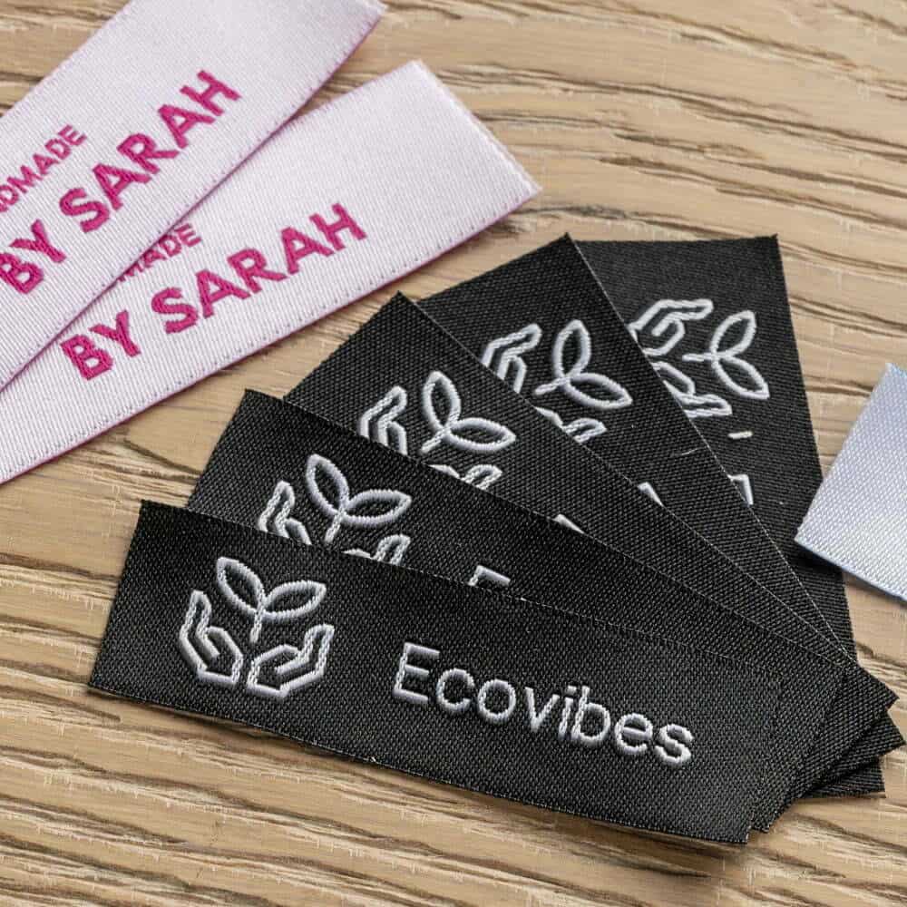 Black Ecovibes woven labels with white hands-and-leaves icons on wood beside pale pink Handmade by Sarah strips.