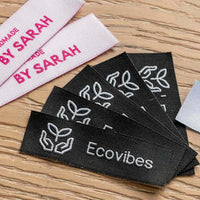 Black Ecovibes woven labels with white hands-and-leaves icons on wood beside pale pink Handmade by Sarah strips.