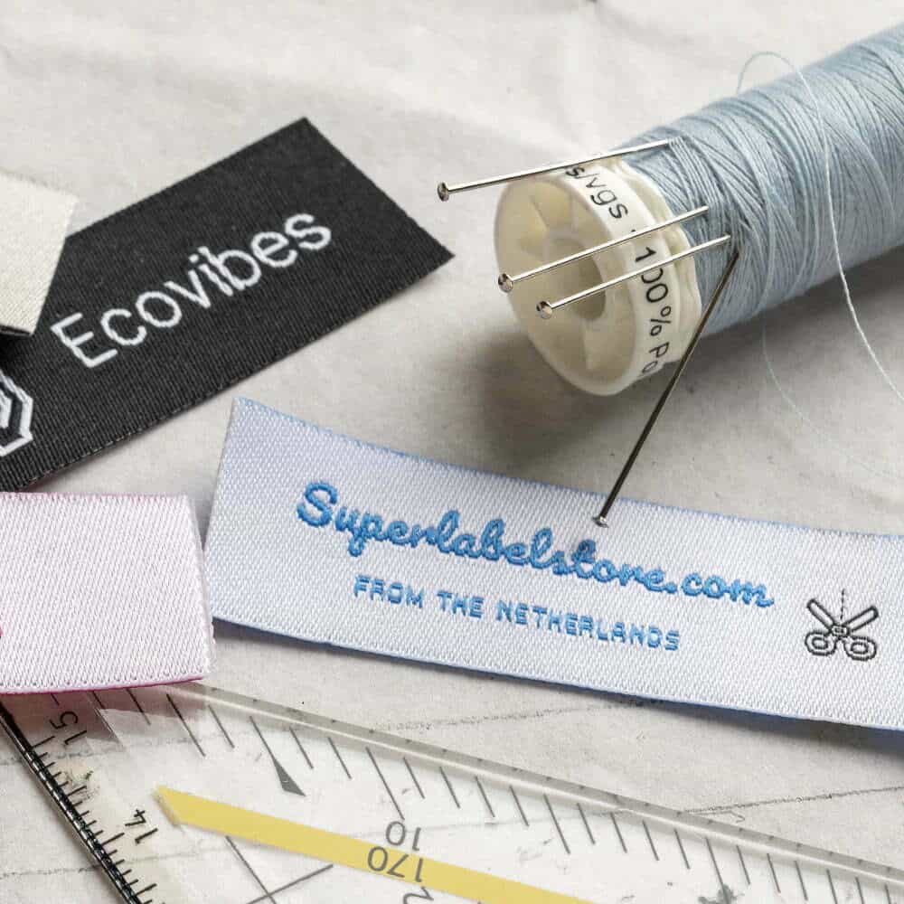 White fabric label with blue Superlabelstore.com script and a black scissors icon beside thread, pins and a ruler.