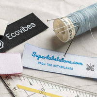 White fabric label with blue Superlabelstore.com script and a black scissors icon beside thread, pins and a ruler.