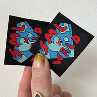 Two black square fabric labels held in a hand, with blue and mint graphics and red lettering woven into the surface.