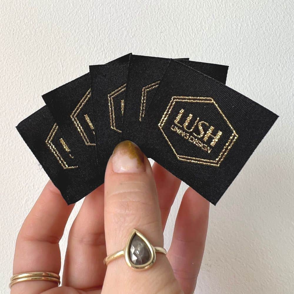 Black square woven labels fanned in a hand, showing gold-toned LUSH lettering inside a double hexagon outline.