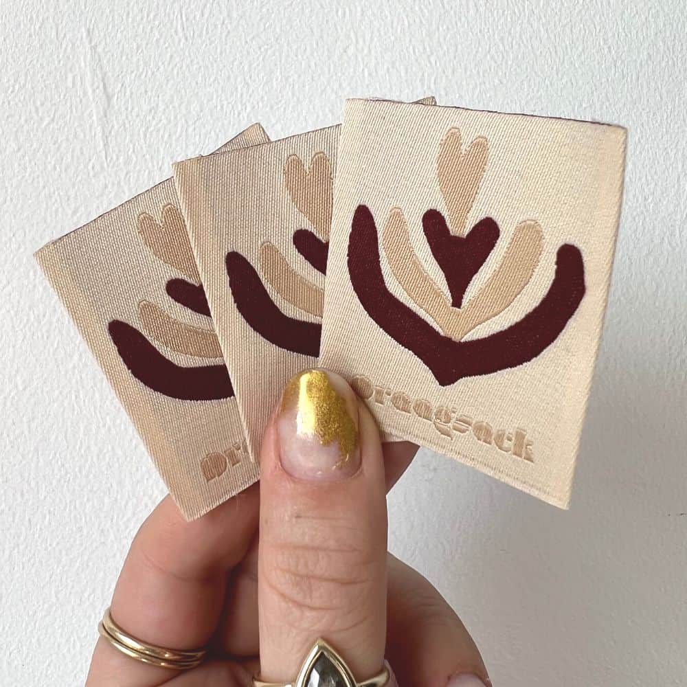 Three cream square woven labels held in a hand, with burgundy and beige heart-shaped abstract motifs.