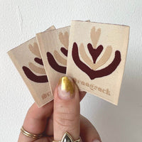 Three cream square woven labels held in a hand, with burgundy and beige heart-shaped abstract motifs.