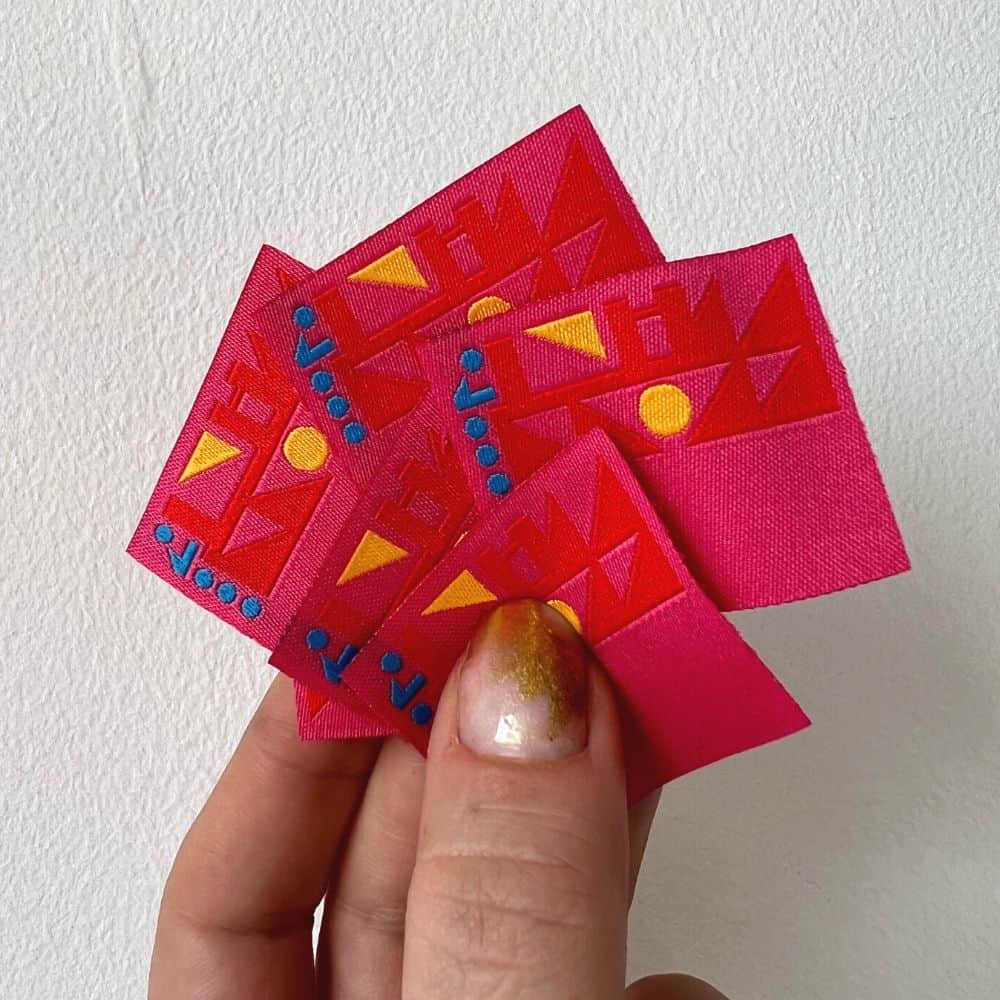 Bright pink woven squares held in a hand, with red geometric shapes, yellow triangles and circles, and blue dots.
