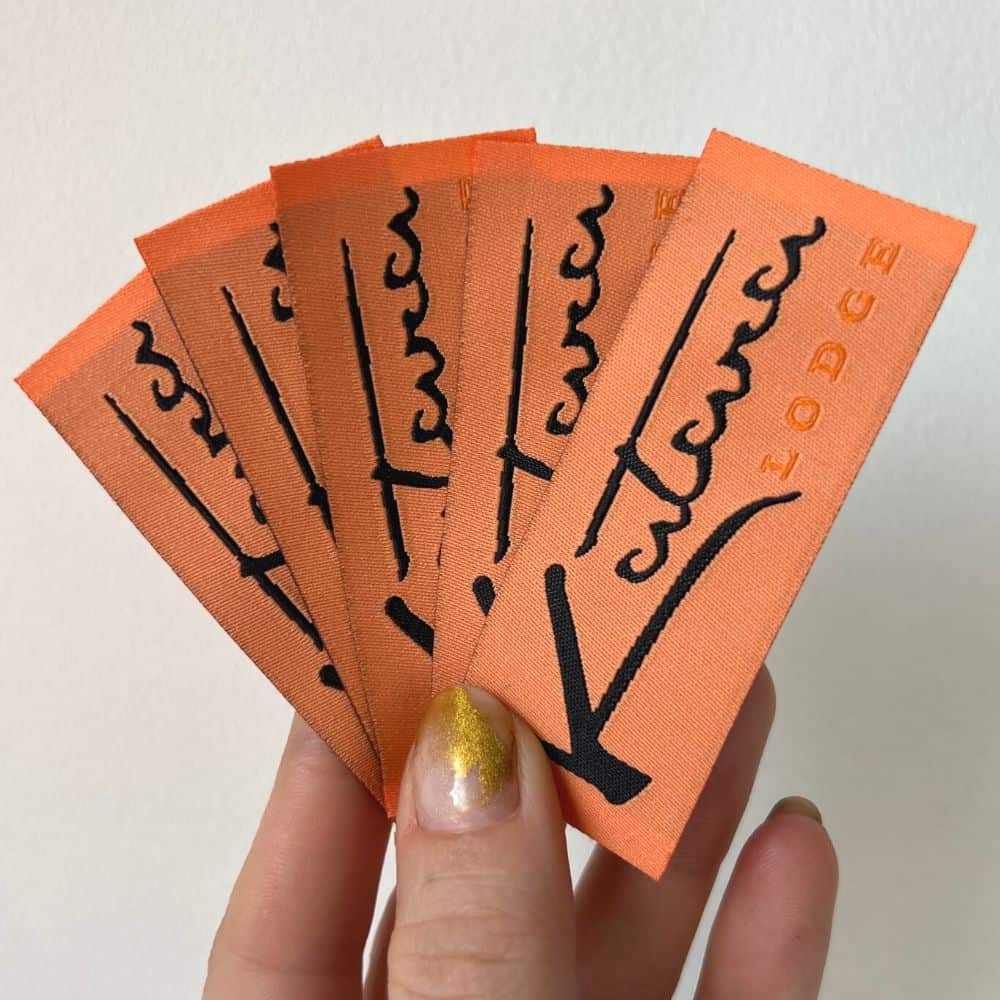 Orange fabric labels fanned in a hand, showing black script lettering and a fine woven texture.