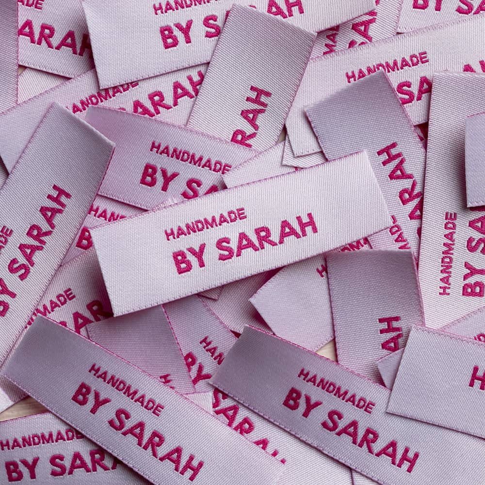 A pile of pale pink rectangular woven labels with magenta Handmade by Sarah lettering and visible thread texture.