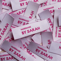 A pile of pale pink rectangular woven labels with magenta Handmade by Sarah lettering and visible thread texture.