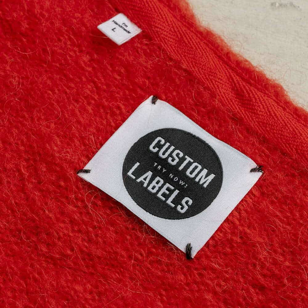 White square woven label with a black Custom Labels badge, stitched at four corners onto red knitwear.