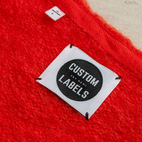 White square woven label with a black Custom Labels badge, stitched at four corners onto red knitwear.