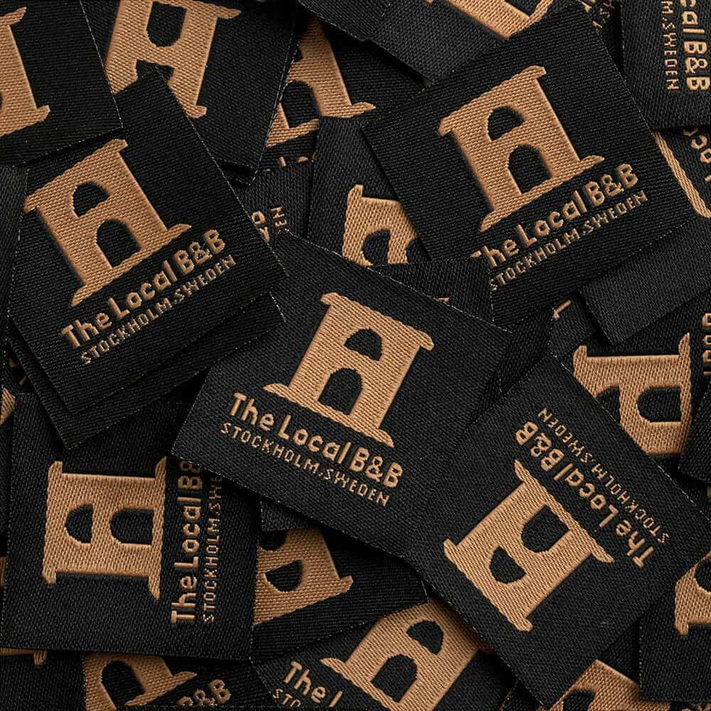 Overlapping black woven labels with tan building emblems and The Local B&B Stockholm Sweden lettering.