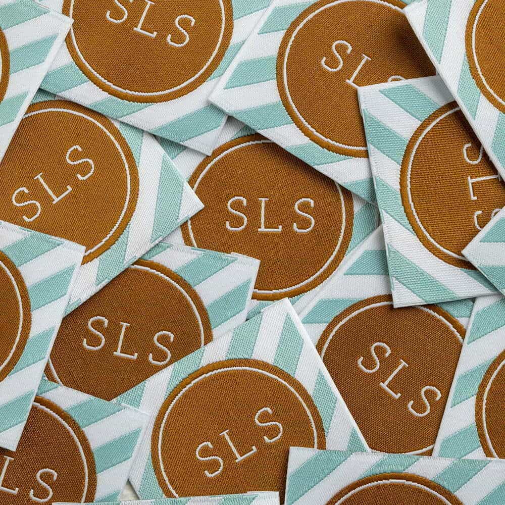 Square woven labels in a pile, each with a brown SLS medallion bordered by mint and white diagonal stripes.
