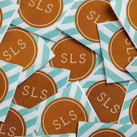 Square woven labels in a pile, each with a brown SLS medallion bordered by mint and white diagonal stripes.