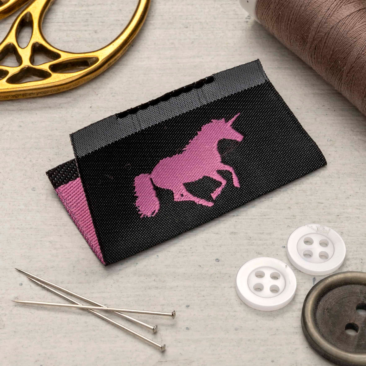 Folded black woven tag with a pink unicorn silhouette, displayed beside pins, white buttons and a spool of thread.